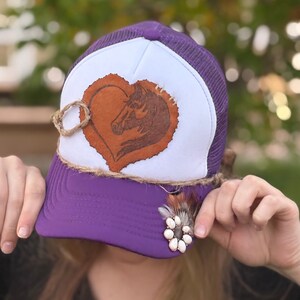 May include: Purple and white trucker hat with a brown leather heart featuring a horse silhouette. The hat has a brown braided trim and a white feather with a turquoise stone embellishment.