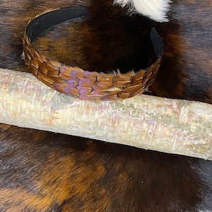 May include: A brown feather headband with purple accents. The headband is on a brown and white cowhide background.