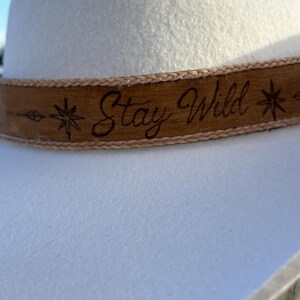 May include: A white felt hat featuring a tan leather band wrapped around the base. The leather band is engraved with the text "Stay Wild" in a cursive script, flanked by two star-shaped symbols and a small arrow design on the left side.