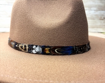 Op de afbeelding: Een beige fedora hoed met een brede rand en een ingedeukte kroon. De hoed is versierd met een band van veelkleurige veren in tinten bruin, zwart, wit en iriserend blauw. De tekst "One of a Kind" staat op de voorkant van de kroon gedrukt.