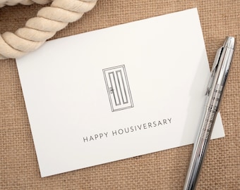 May include: A white rectangular greeting card featuring a black line drawing of a door and the text "HAPPY HOUSIVERSARY" in black capital letters, accompanied by a silver metallic pen with a textured grip and a thick beige rope.