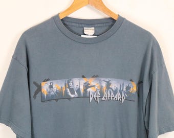 Puede incluir: Una camiseta azul grisáceo de la banda Def Leppard. La camiseta presenta un gráfico rectangular con los miembros de la banda y el nombre de la banda en letras blancas estilizadas. La camiseta está colgada en una percha de madera.