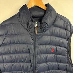 May include: Navy blue quilted vest with a high collar and a full-length zipper. The vest features a small red logo on the chest. The vest is hanging on a wooden hanger.