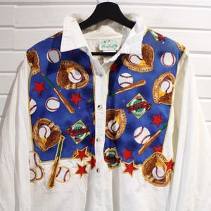 Op de afbeelding: Wit overhemd met baseball-thema. Het shirt heeft een blauw paneel met honkballen, handschoenen, knuppels en sterren. De kraag en mouwen zijn wit en het shirt is dichtgeknoopt.