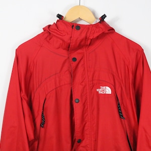 May include: A bright red hooded jacket featuring a front zipper closure with a snap button placket, a high collar, and two zippered pockets. The North Face logo is embroidered in white on the upper left chest area of the garment.