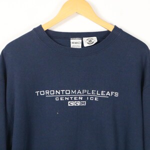May include: A navy blue long-sleeve sweatshirt featuring white embroidered text that reads TORONTO MAPLE LEAFS CENTER ICE with a CCM logo box underneath. The garment has a crew neckline and is displayed on a wooden hanger against a white background.