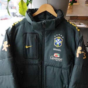 Peut inclure: Une veste matelassée zippée à manches longues vert foncé arborant l'écusson CBF Brasil avec cinq étoiles, un logo Nike jaune et le logo Guaraná Antarctica. Les manches présentent des motifs jaunes et la poitrine inclut une poche zippée verticale.