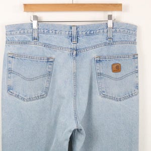 May include: Light blue denim jeans featuring a high-waisted design with belt loops, a button fly, and two back pockets. The right back pocket displays a small, square, brown leather-like patch with the Carhartt brand logo embossed on it.