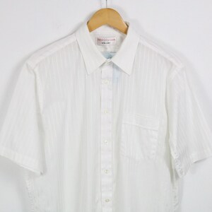 May include: White short-sleeved shirt with vertical stripes, a chest pocket, and a button-down front. The shirt is hanging on a wooden hanger. The label reads "Melody Club".