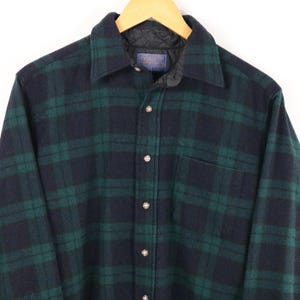 Pode incluir: Uma camisa de manga comprida com botões e um padrão xadrez em azul-marinho escuro e verde-floresta. A camisa tem um colarinho clássico, um bolso no peito e um forro acolchoado preto visível no interior do colarinho com uma etiqueta Pendleton.