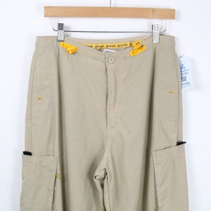 May include: Beige cargo pants featuring a button closure, a yellow drawstring at the waist, and side cargo pockets. The pants include small yellow accents on the pockets and a yellow interior waistband lining with repeating text patterns.
