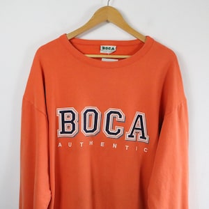 May include: An orange long-sleeve crewneck sweatshirt featuring the word BOCA in large navy blue and white block letters across the chest, with the word AUTHENTIC printed in smaller white letters underneath. The garment hangs on a wooden hanger.