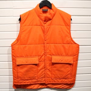 May include: Bright orange quilted vest with a stand-up collar and two front pockets. The vest is hanging on a black hanger against a white slatted background. The vest has a button closure and a quilted design.