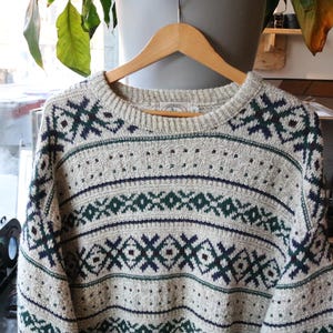 May include: A long-sleeve crew neck sweater in a light oatmeal color featuring a repeating geometric pattern in dark navy blue and forest green. The knit garment has a ribbed collar, cuffs, and hem, with horizontal bands of intricate diamond and cross motifs.