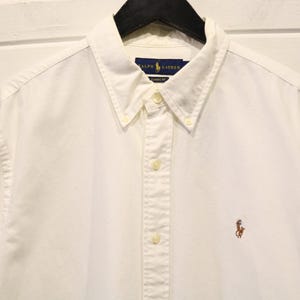 May include: A white button-down shirt with long sleeves, a button-down collar, and a small embroidered logo of a polo player on the chest. The shirt is made of a soft, light-colored fabric and is hanging on a black hanger.