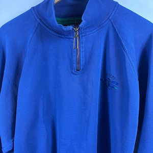 May include: A bright blue quarter-zip pullover sweatshirt featuring a high collar with a metal zip pull. The garment displays a small embroidered graphic on the left chest area with blue and green details and text above the design.