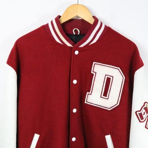 Peut inclure: Une veste universitaire bordeaux avec des manches blanches, arborant un grand écusson en forme de lettre D blanche sur la poitrine et un petit écusson sur la manche représentant deux drapeaux croisés, dont le drapeau canadien. Col rayé et boutons blancs.