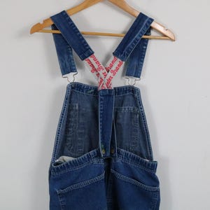 Könnte beinhalten: Blaue Jeans-Latzhose mit verstellbaren Trägern. Die Träger haben einen roten Text mit der Aufschrift "Water Brand". Die Latzhose hängt an einem Holzbügel vor weißem Hintergrund. Die Latzhose hat Taschen und Metallbeschläge.