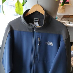 May include: A two-toned zip-up jacket featuring a dark grey upper section and a blue lower section. The jacket has a high collar, a vertical zippered chest pocket, and the white The North Face logo embroidered on the left chest area.