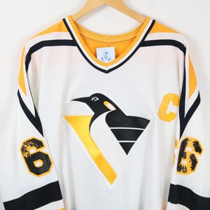 Op de afbeelding: Een wit, zwart en geel ijshockey-shirt met een grote pinguïngrafiek op de borst. De mouwen tonen het nummer 66 in het zwart met gele omlijningen, en een gele letter C bevindt zich op het bovenste borstgedeelte. De V-hals is geel van kleur.
