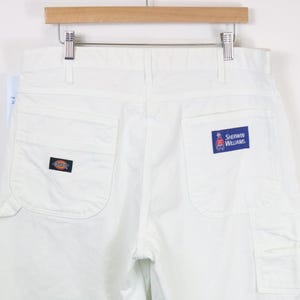 May include: White Dickies work pants featuring a back pocket with a small Dickies logo patch and another back pocket with a blue Sherwin Williams rectangular patch. The pants are hanging from a wooden hanger against a plain white background.