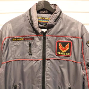Puede incluir: Chaqueta gris con ribetes rojos y cremallera negra. Cuenta con un parche "TRANS AM" con un emblema de ave rojo y dorado. Incluye texto "STYLE AUTO" y un pequeño bolsillo con cremallera. Chaqueta de estilo vintage.