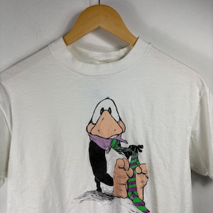 Pode incluir: Camiseta branca com um personagem de desenho animado de pato com um cachecol roxo e uma gravata listrada verde e roxa. O pato é preto e branco com um bico rosa. A camiseta está em um cabide de madeira.