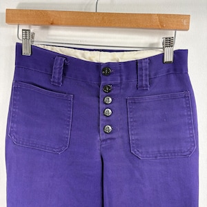 Könnte beinhalten: Nahaufnahme einer lila Hose mit zwei Vordertaschen und einem Knopfverschluss. Die Hose hängt an einem Holzbügel. Die Knöpfe sind schwarz und die Hose hat einen weißen Innenbund.