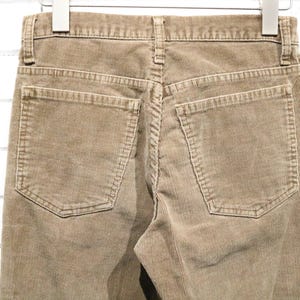 May include: Close-up of tan corduroy trousers hanging on a hanger. The trousers have two back pockets and a classic five-pocket design. The corduroy fabric has a ribbed texture.