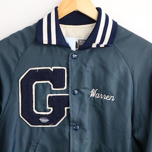 May include: A teal blue varsity jacket with a white and navy blue striped collar. The jacket features a large navy blue letter "G" with a white outline and the name "Warren" embroidered in white.