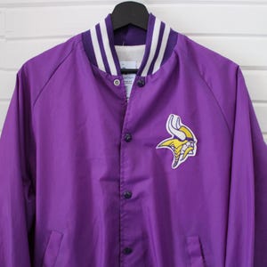 May include: Purple satin jacket with a snap-button front and a white and purple striped collar. The jacket features a team logo patch on the chest. The jacket is hanging on a black hanger.