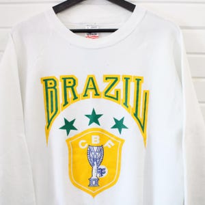 Puede incluir: Sudadera blanca con la palabra "BRAZIL" en letras verdes y amarillas. Debajo, tres estrellas verdes y un escudo amarillo con un trofeo y las letras "CBA".