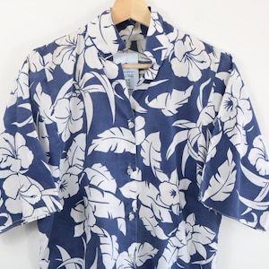 May include: A short-sleeved button-down shirt featuring a dark blue background with a repeating white floral and leaf pattern. The garment has a classic collar and a front button closure, displayed on a wooden hanger against a plain background.
