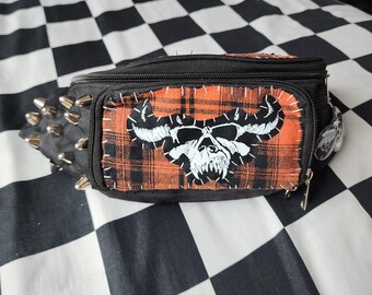 May include: A black bum bag featuring a rectangular orange and black plaid patch with a white screen-printed horned skull graphic. The bag includes a zippered main compartment and silver-toned metal cone studs along the side panel.