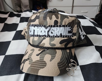 May include: A camouflage baseball cap featuring a black and white patch with the text UNHOLY GRAVE stitched onto the front. The cap includes a black cord trim across the brim and three silver-toned metal rings attached to the side of the visor.