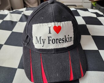 May include: A black baseball cap featuring a rectangular white fabric patch sewn onto the front panel. The patch displays the text I, a red heart symbol, and the words My Foreskin. The brim has three vertical red fabric accents.