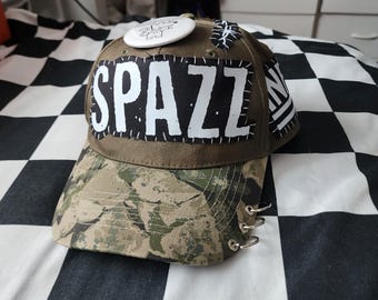 May include: An olive green baseball cap featuring a black rectangular patch with the white text SPAZZ stitched onto the front. The brim has a camouflage pattern with three silver metal rings attached to the side. A white circular pin is on top.