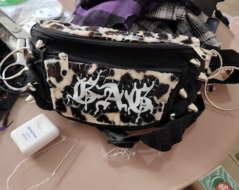 May include: A black fanny pack featuring a leopard print faux fur panel on the front with a white embroidered patch displaying the text GAG. The bag is accented with silver metal spikes and large circular rings on the sides, with a black adjustable strap.