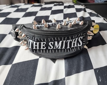 May include: A black bum bag featuring silver metal spikes along the top edge and a rectangular patch on the front with the white text THE SMITHS. The bag has a zippered closure and a yellow circular pin attached to the side strap area.