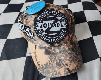 May include: A camouflage baseball cap featuring a circular black and white patch with the text ZOUNDS and CAN'T CHEAT KARMA WAR-SURVIVOR. The cap includes a bright blue circular pin, a black and white rectangular patch, and several silver safety pins.