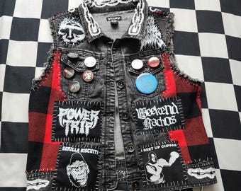 May include: A sleeveless denim waistcoat with a black and red buffalo plaid pattern on the sides. The garment features various band patches including Power Trip, Weekend Nachos, and Gorilla Biscuits, along with several circular pinback buttons attached.