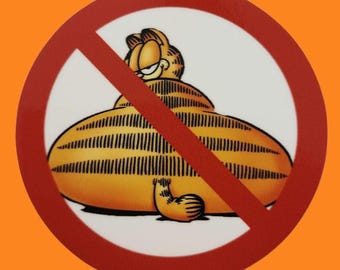May include: A circular sticker featuring Garfield, the cartoon cat, in a large, round form, crossed out with a red diagonal line. The cat is orange with black stripes, set against a white background, with an orange border.