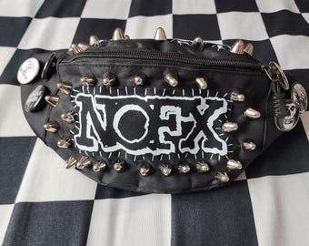 May include: A black bum bag featuring a white NOFX patch stitched onto the front. The bag is decorated with numerous silver metal spikes along the top and bottom edges, and several circular pinback buttons attached to the sides and strap.