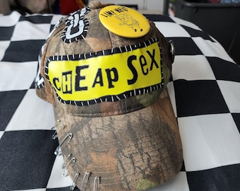 May include: A camouflage baseball cap featuring a yellow rectangular patch with the black text CHEAP SEX stitched onto the front. The cap is decorated with multiple silver safety pins along the brim and a yellow circular button with a cartoon character.