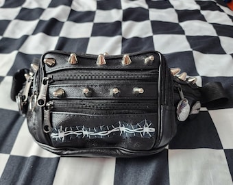 May include: A black fanny pack featuring silver-toned metal spikes along the top and front panels. The bag includes multiple zippered compartments and a white graphic print of barbed wire across the lower front section with a black strap.