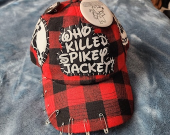May include: A red and black plaid baseball cap featuring a white patch with the text WHO KILLED SPIKEY JACKET? in black. The cap is decorated with several silver safety pins along the brim and two circular pinback buttons on the crown.