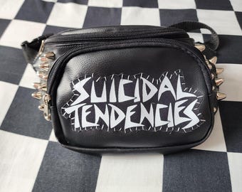 May include: A black faux leather bum bag featuring a white Suicidal Tendencies patch stitched onto the front pocket. The bag is accented with silver-toned metal spikes along the sides and includes a black zip closure and an adjustable strap.