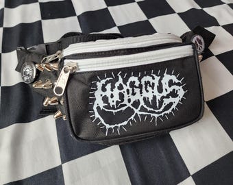 May include: A black bum bag featuring a white zip and a white patch with the text HAGGUS in a stylised font. The bag is adorned with silver-toned metal spikes along the side and a circular pin with a skull graphic attached to the strap.