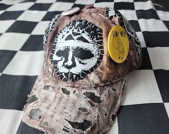 May include: A distressed brown camouflage baseball cap featuring a large black and white graphic of a skull on the front panel. The cap is adorned with a yellow circular pin displaying a cartoon character and the text Limp Moist.