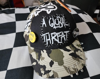May include: A black and camouflage baseball cap featuring a white patch with the text A GLOBAL THREAT, a yellow circular pin reading SPAZZ, a black circular pin reading DISRUPT, and several silver safety pins attached to the brim and side.